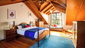 Un dormitorio con techo de madera, una cama grande con manta azul, una mesita de noche, un aparador y una ventana con cortinas.