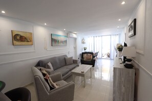 A modern living room with a grey sofa, a coffee table, and a TV. There are paintings on the walls and a window with curtains.
