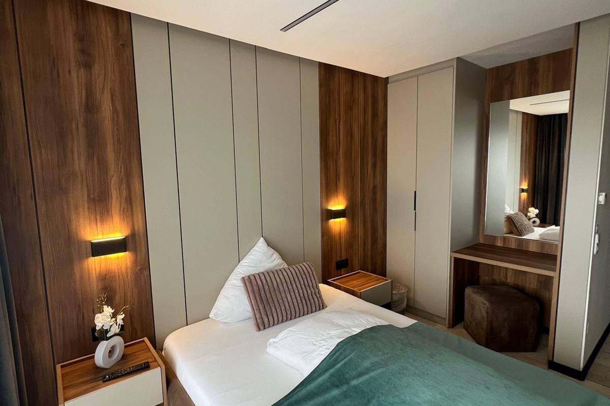 A modern bedroom with a large bed, wooden panel walls, a nightstand with a lamp, and a mirror.