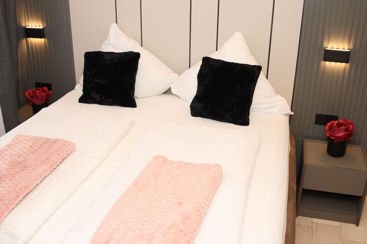 A neatly made bed with white and black pillows, a pink throw blanket, and a red rose on a bedside table.