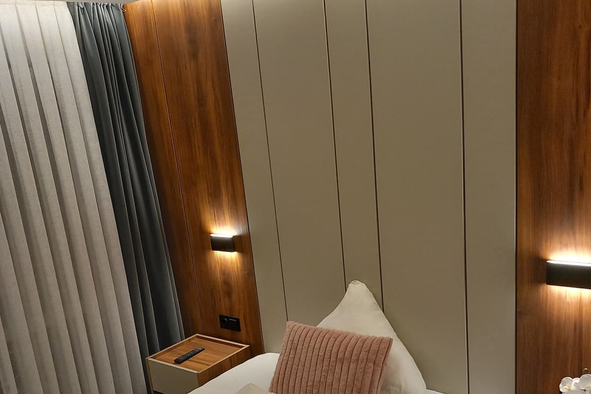 A modern hotel room with a large bed, wooden panel walls, a nightstand with a lamp, and a window with curtains.