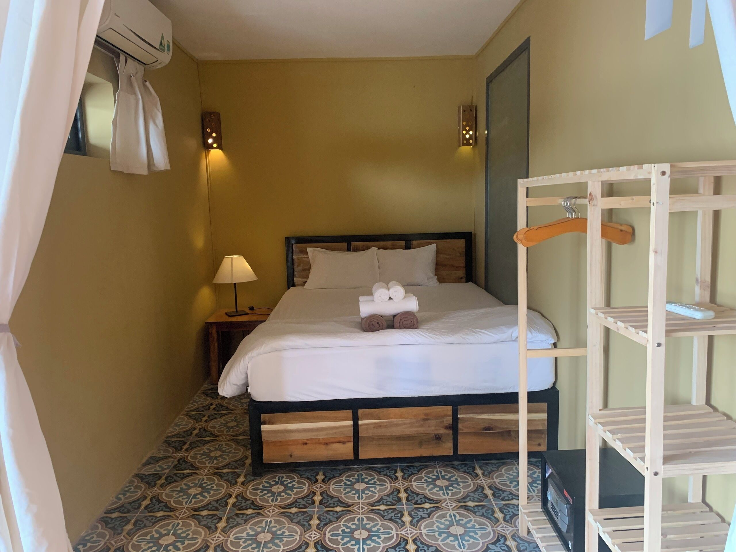 A small, cozy room with a bed, a nightstand, a lamp, a wooden bunk bed, and a tiled floor.