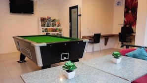 A pool table with a green felt surface, a bookshelf with various items, a door, a small table with two potted plants, and a red couch. - Royal Waves Hotel (Patong)