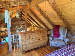 A wooden interior with a high ceiling, a wooden dresser, a patterned armchair, and a bed with a plaid bedspread.