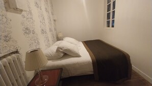 Un dormitorio pequeño y ordenado, con cama, lámpara de noche, radiador y ventana con vistas.