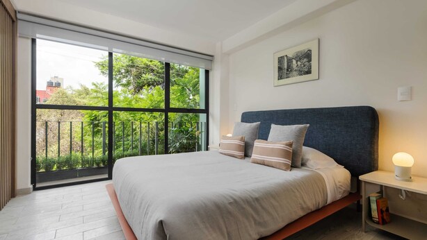 A bedroom with a large window, a bed with pillows, a bedside table with a lamp, and a framed picture on the wall.
