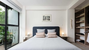 Une chambre moderne avec un grand lit, deux tables de chevet avec des lampes, un tableau encadré au mur et une vue sur la verdure par une porte coulissante.