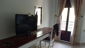 Ein Zimmer mit Fernseher auf einem Holztisch, einem Sessel, einem Fenster mit Vorhängen und Blick ins Freie.
