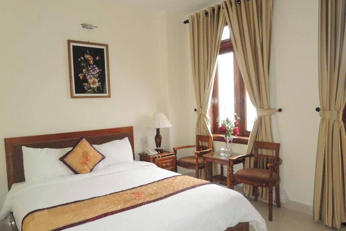 A hotel room with a bed, a desk, chairs, a window with curtains, and a wall-mounted picture.