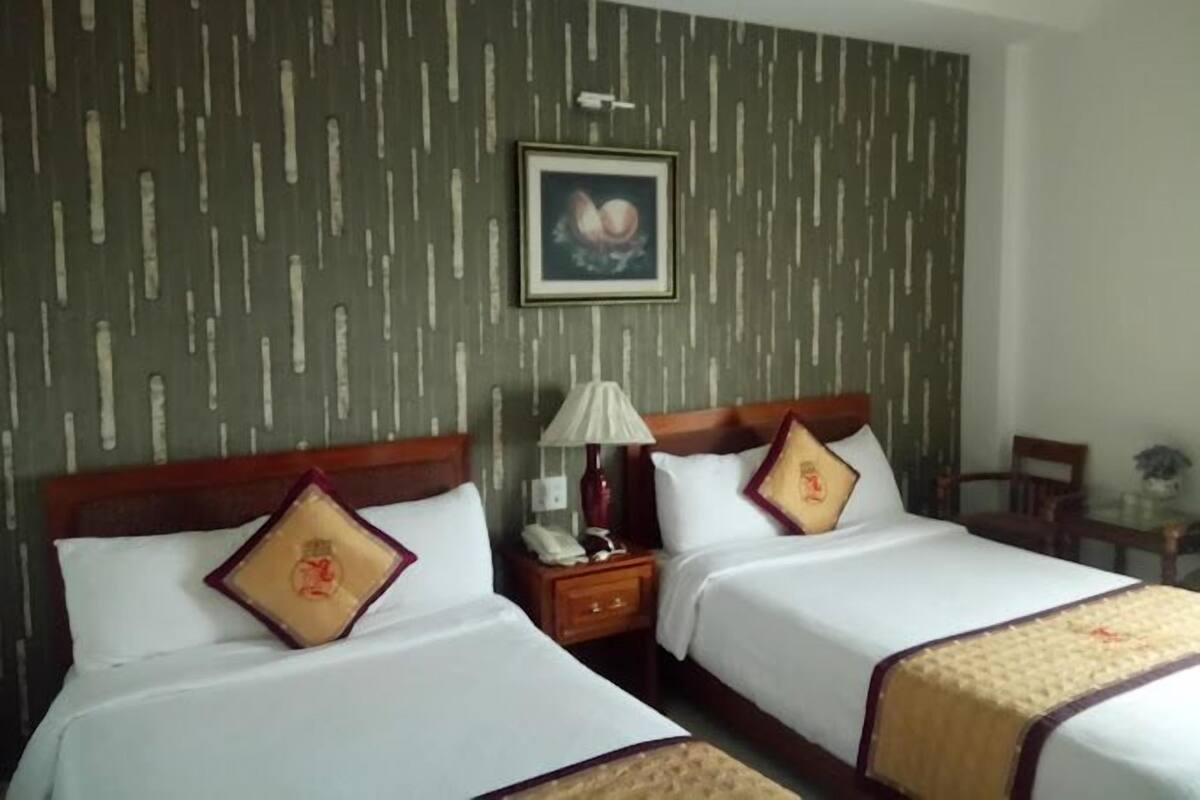 A hotel room with two beds, a nightstand with a lamp, a framed picture on the wall, and a small table with a chair.