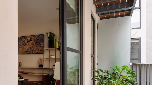 Ein Balkon mit Holzboden, einer Topfpflanze und einer Schiebetür zu einem Innenraum mit Schreibtisch, Stuhl und Regalen.