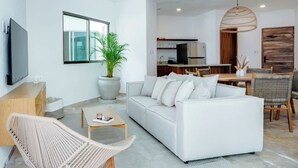 Ein modernes Wohnzimmer mit einem weißen Sofa, einem Couchtisch, einem Korbstuhl, einer großen Topfpflanze und einem an der Wand befestigten Fernseher.