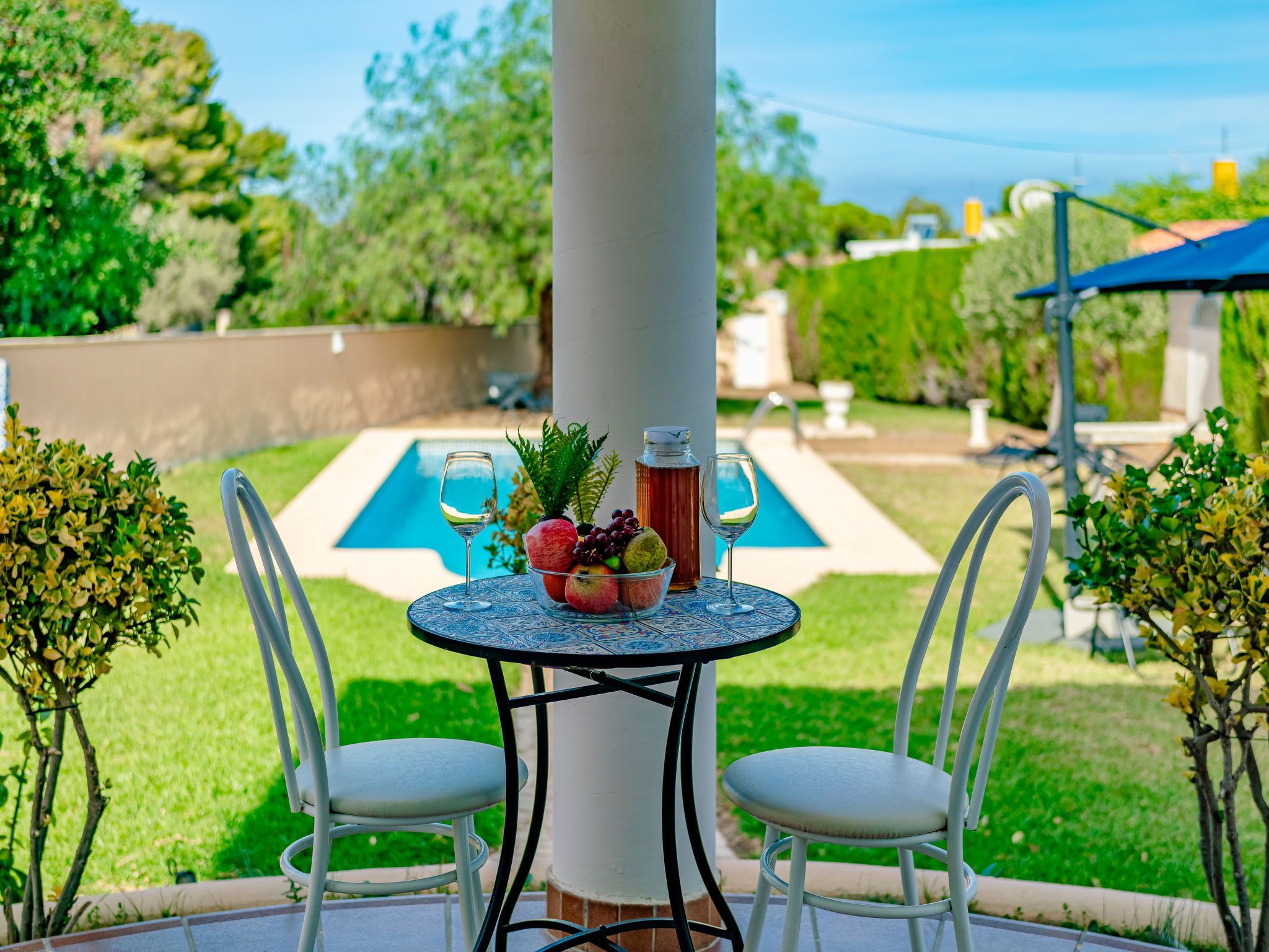 Um pátio com uma mesa redonda, duas cadeiras brancas, uma cesta de frutas e dois copos de vinho.