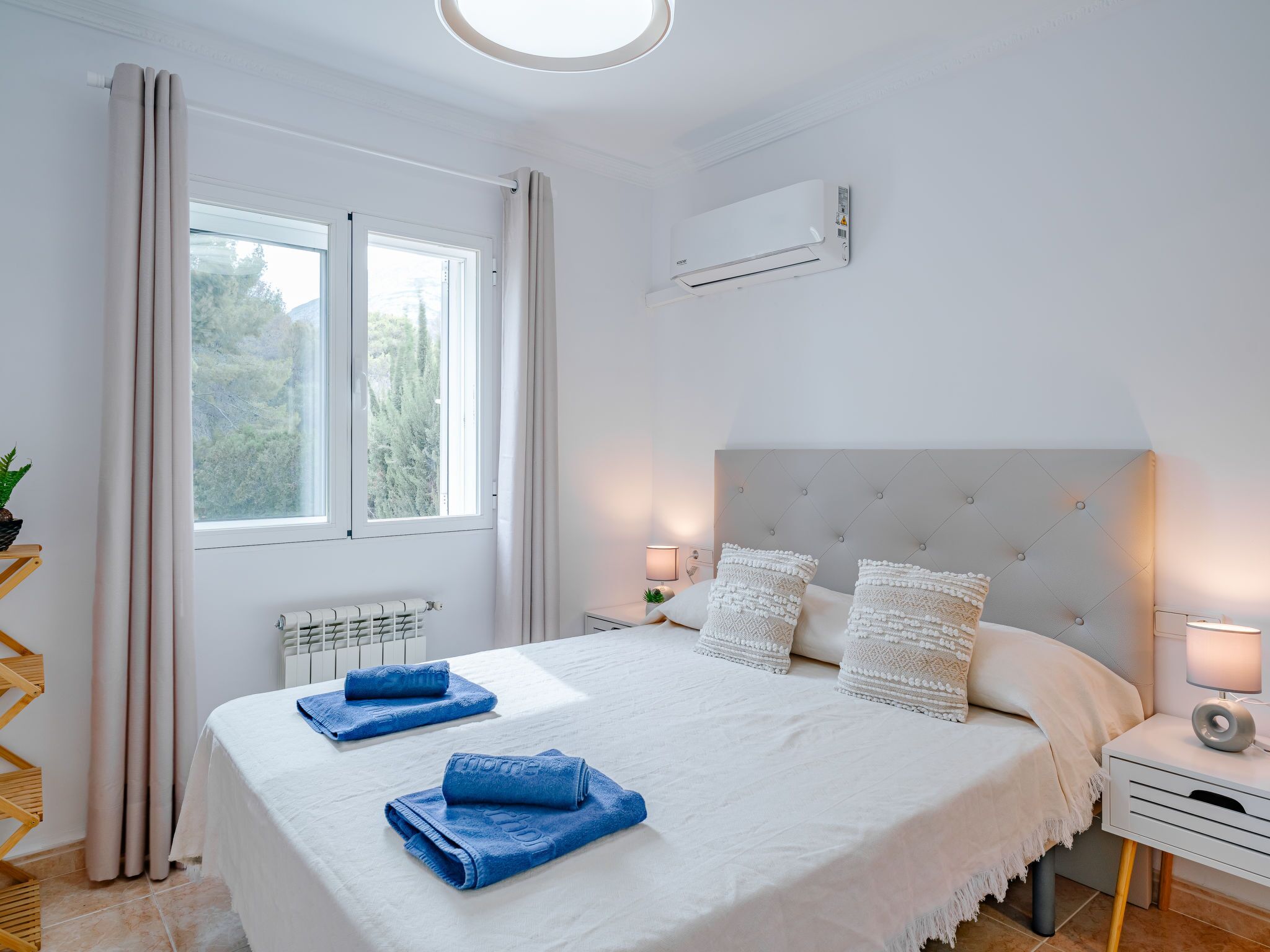 Ein Schlafzimmer mit einem großen Bett, einem Heizkörper, einem Fenster mit Vorhängen, einem Nachttisch mit Lampe und einer Klimaanlage.