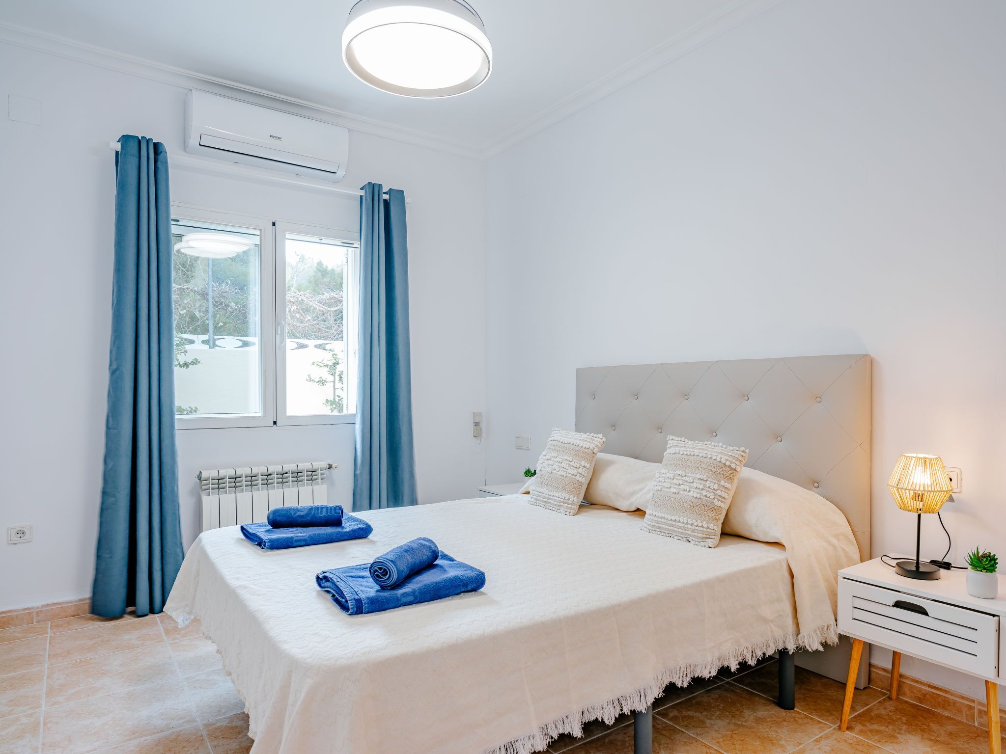A neatly arranged bedroom with a bed, a bedside table with a lamp, and a window with curtains.