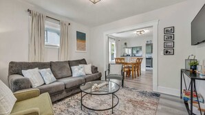 Ein Wohnzimmer mit grauem Sofa, einem runden Glastisch und einem gemusterten Teppich. Durch einen offenen Durchgang ist ein Essbereich mit Holztisch und Stühlen zu sehen.