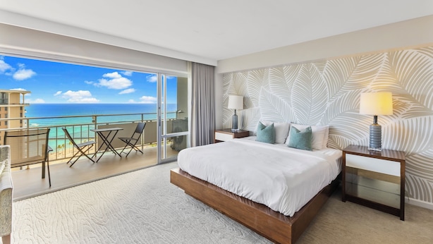 A hotel room with a large bed, a nightstand, a lamp, a view of the ocean, and a balcony with chairs and a table. - Ilikai Hotel Studio (Honolulu)