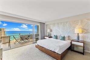 Hotelkamer met een groot bed, nachtkastje, lamp, uitzicht op de oceaan en een balkon met stoelen en een tafel.
