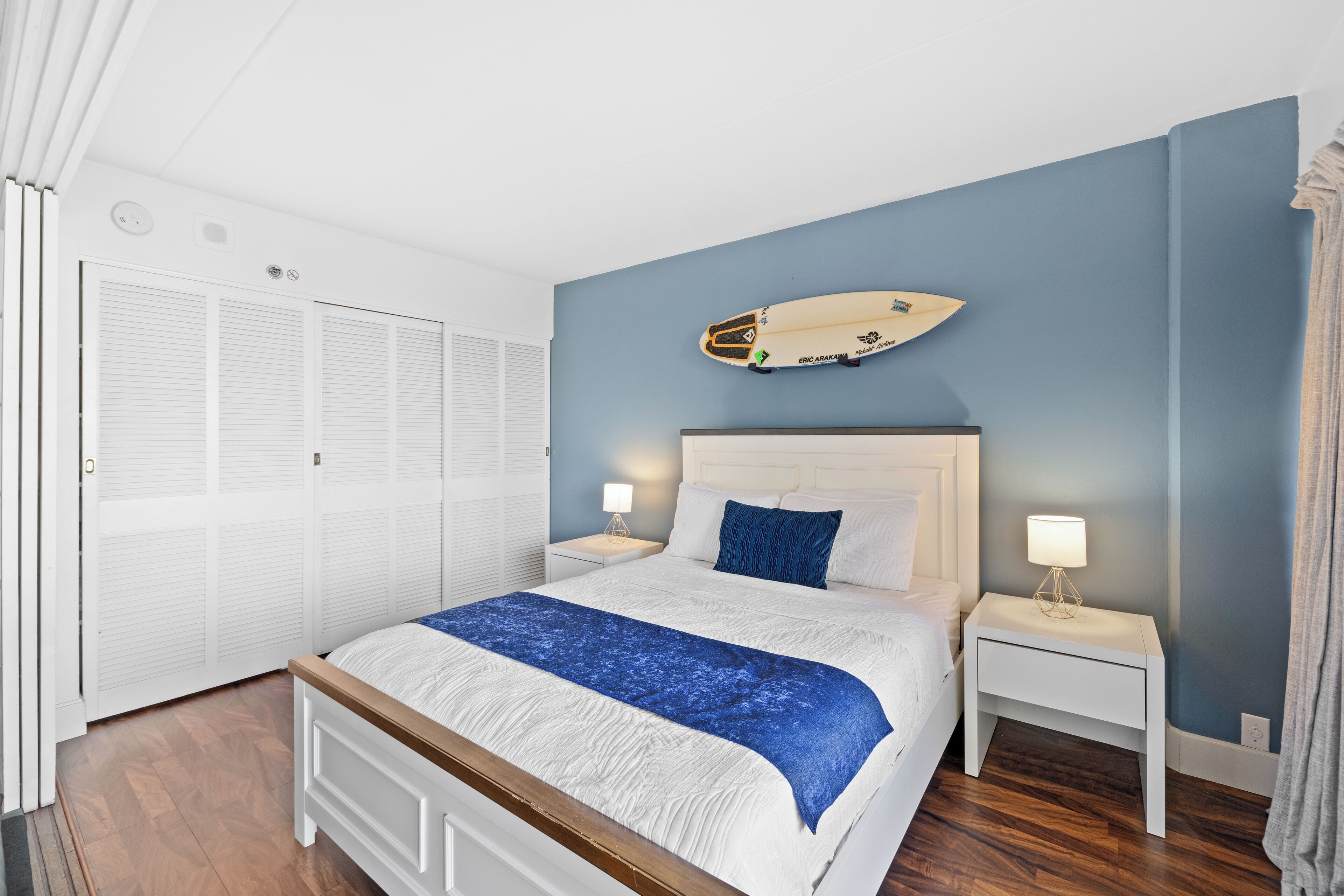 A bedroom with a bed, white nightstands, a surfboard on the wall, and a closet with white shutters.