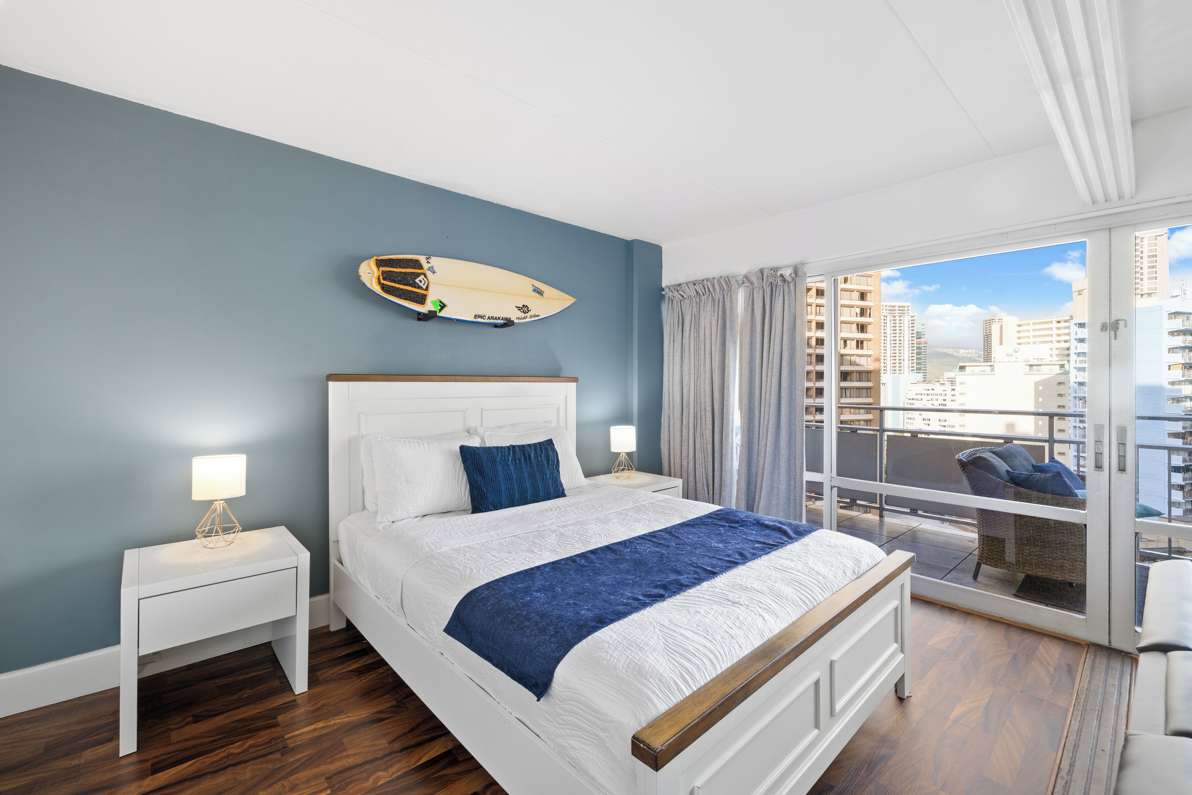 A bedroom with a bed, nightstands, a surfboard on the wall, and a balcony with a view of buildings.