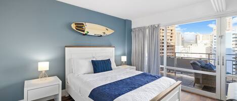 A bedroom with a bed, nightstands, a surfboard on the wall, and a balcony with a view of buildings.