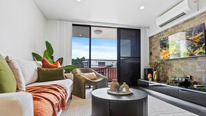 Un salon moderne comprenant un canapé, un fauteuil et une table basse, une grande fenêtre donnant sur la ville et un mur décoratif en pierre agrémenté d’un tableau.