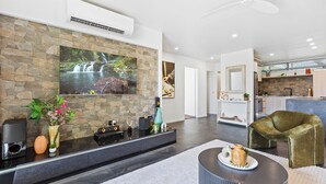 Un salon moderne avec un mur en pierre, un téléviseur à écran plat, un meuble de rangement noir, un fauteuil en velours vert et une cuisine en arrière-plan.