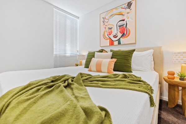 A neatly made bed with a green blanket, a pillow with a striped pattern, and a green pillow. There is a small table with a plant and a candle. A window with blinds is visible, and a colorful painting hangs on the wall.