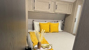 Un lit bien fait, avec un plaid jaune et des coussins, un vase contenant des plantes séchées et des armoires en bois au-dessus.