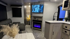 Un salon moderne comprenant un canapé, un téléviseur fixé au mur, une cheminée et un coin cuisine avec des placards et un plan de travail.