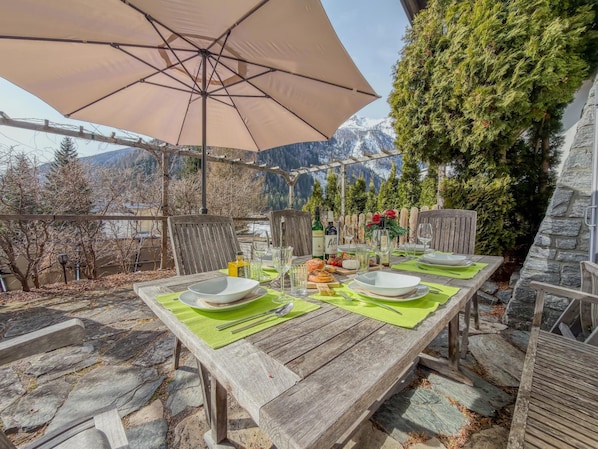 Un espace extérieur aménagé pour les repas, avec une table en bois, des sets de table verts et un grand parasol.