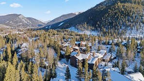 Un village de montagne enneigé, avec des maisons en bois entourées de forêts denses et de collines recouvertes de neige.