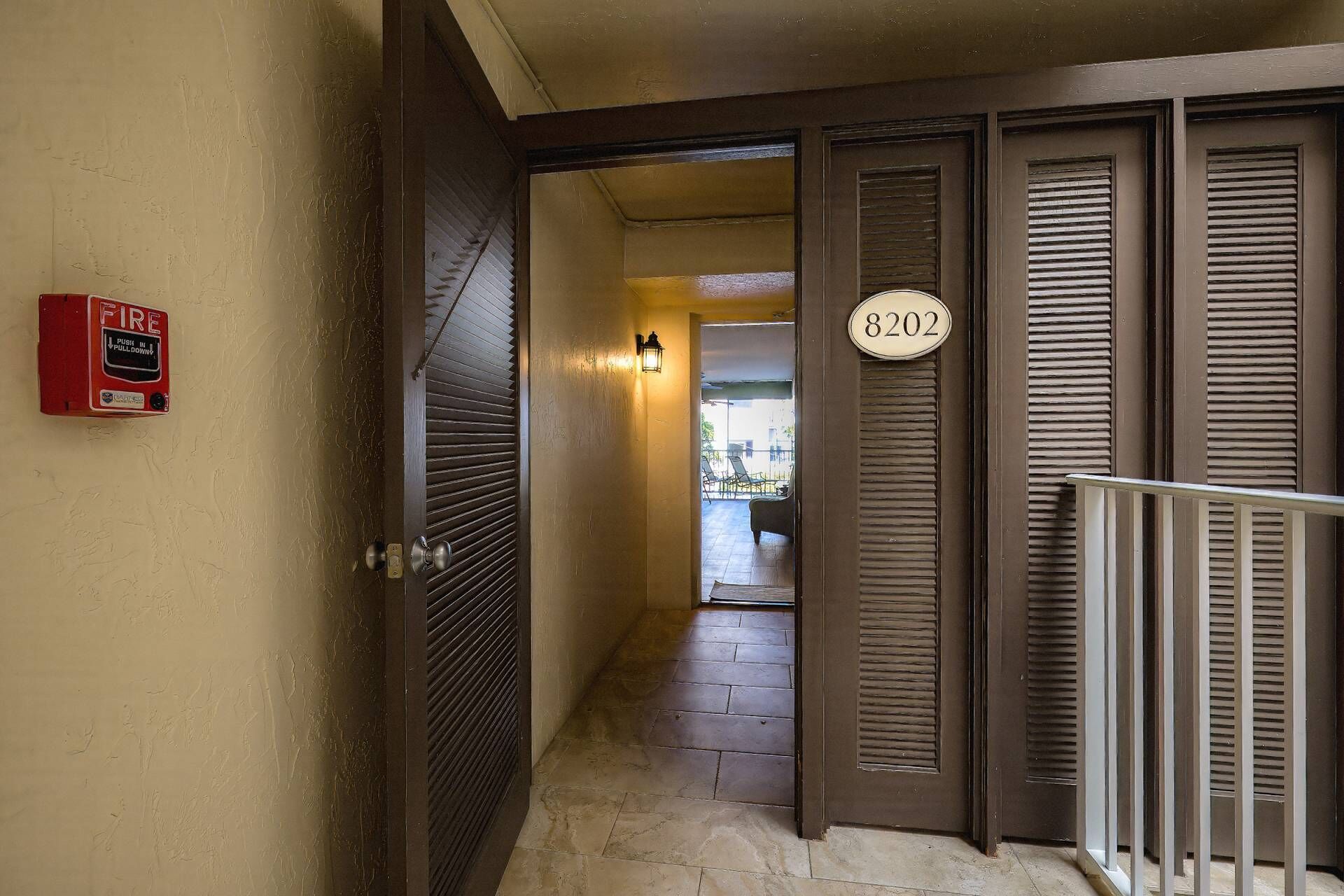 A hallway with a fire alarm, a door with a number plate, and a balcony railing.
