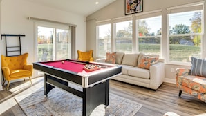 Een pooltafel in een goed verlichte kamer met grote ramen, een gele fauteuil en een witte bank met kleurrijke kussens.