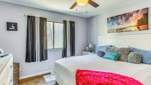 A bedroom with a bed, a ceiling fan, a window with curtains, a painting on the wall, and a nightstand with a lamp. - Walk to Ocean: Myrtle Beach Townhome w/ Patio (Myrtle Beach)