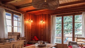 Interior de una cabaña de madera con una mesa de comedor, sillas, un banco y una ventana con vista a los árboles.