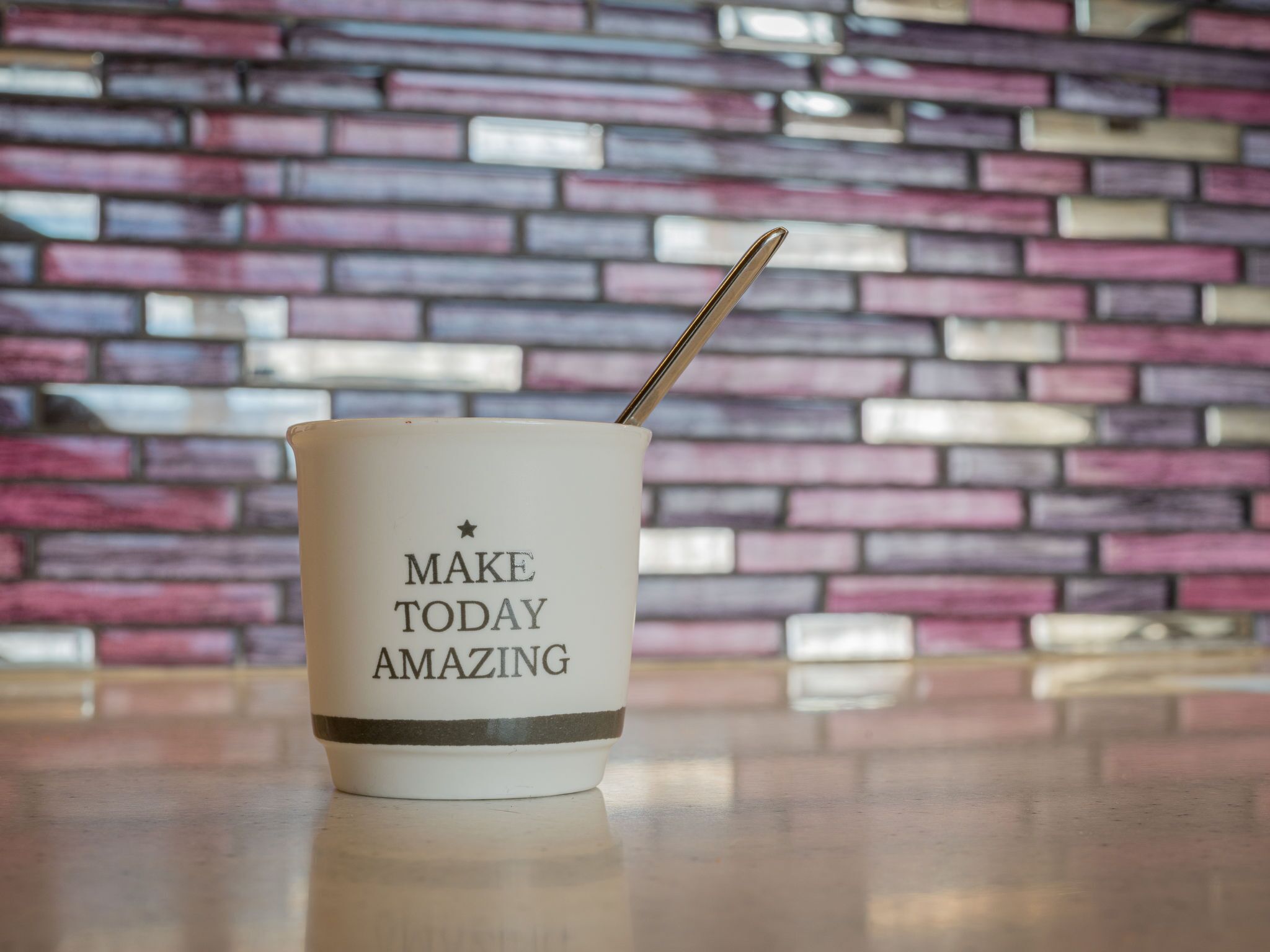 Een witte mok met de tekst 'MAKE TODAY AMAZING' en een lepel erin, geplaatst op een reflecterend oppervlak, met een bakstenen muur op de achtergrond.