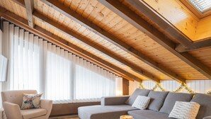 A cozy loft bedroom with a sloped ceiling, wooden beams, a grey sofa, a small white table, and a chair.