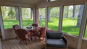 Una terraza techada con una mesa y sillas, un sofá y ventanas amplias que dan a un área verde con árboles.