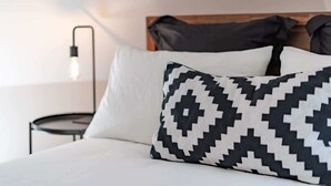 Una cama con un almohadón blanco y otro almohadón con estampado blanco y negro, un cabecero de madera y una mesita de noche con una lámpara.