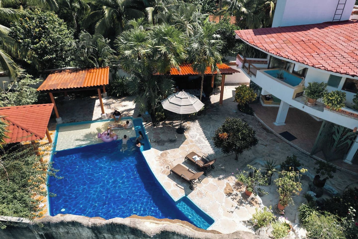 A swimming pool with a sun terrace, surrounded by palm trees and a building with a red-tiled roof.