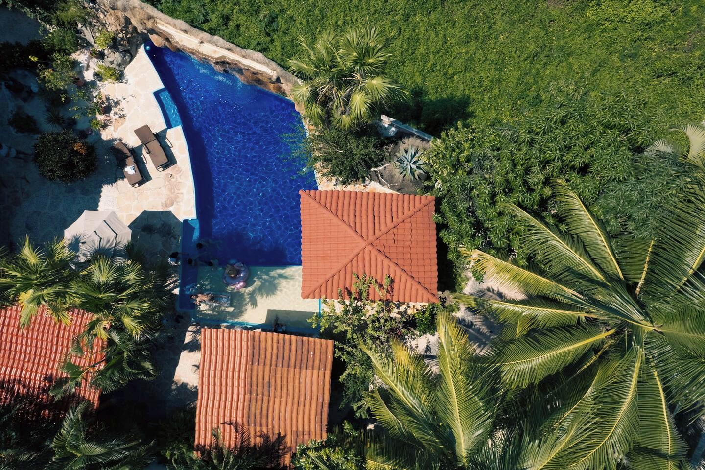 An aerial view of a resort with a swimming pool, palm trees, and a red-roofed building.