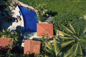Vista aèria d'un complex turístic amb piscina, palmeres i un edifici amb teulada vermella.