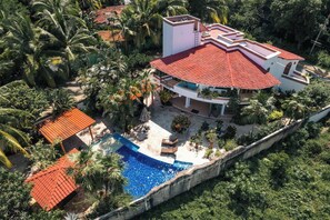 Un edificio de varios pisos con techo de tejas rojas, una piscina y abundante vegetación alrededor de la propiedad.