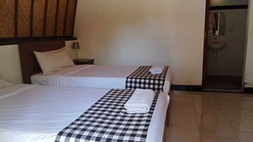 A hotel room with two beds, each with a checkered bedspread and a folded towel. There is a wooden headboard on the left bed, a nightstand with a lamp, and a bathroom visible through an open door.