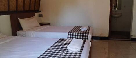 A hotel room with two beds, each with a checkered bedspread and a folded towel. There is a wooden headboard on the left bed, a nightstand with a lamp, and a bathroom visible through an open door.