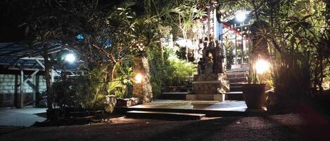 A nighttime view of a garden with a stone statue, surrounded by lush greenery and illuminated by soft lighting.