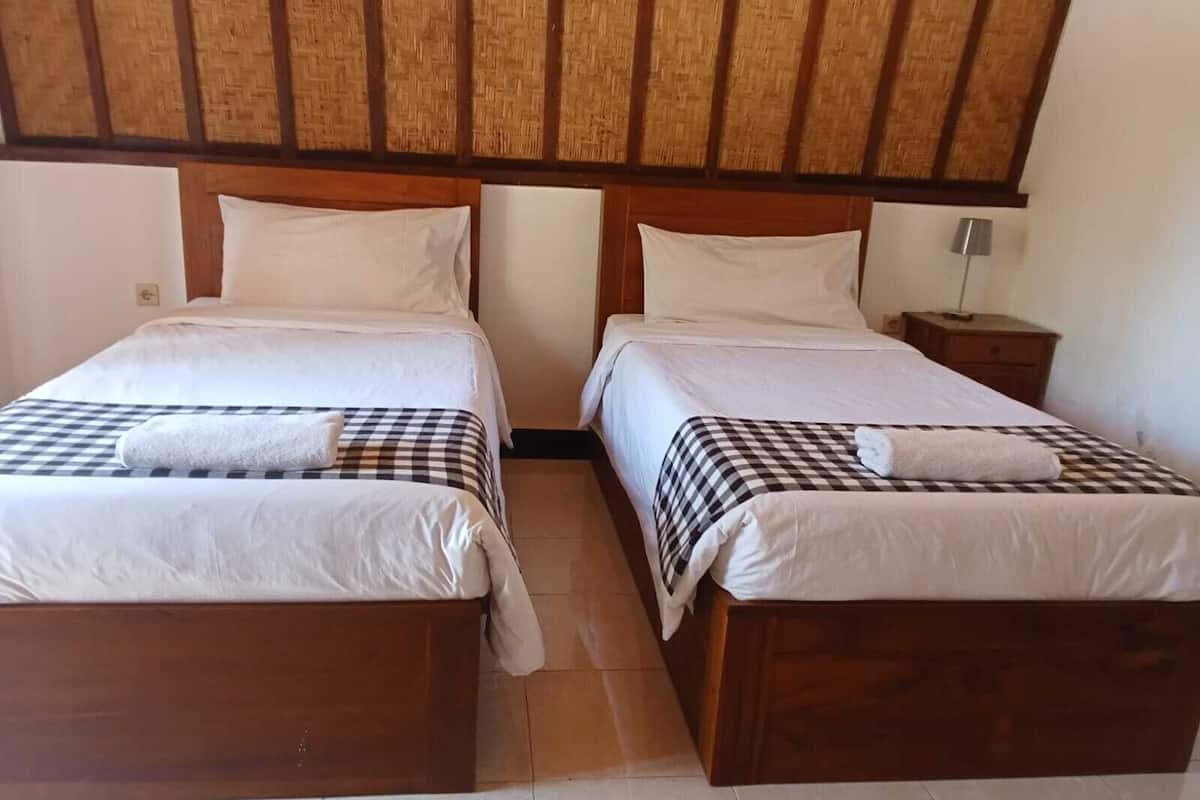 A room with two wooden beds, each with a checkered bedspread and white pillows. There is a small wooden nightstand with a lamp on one side.