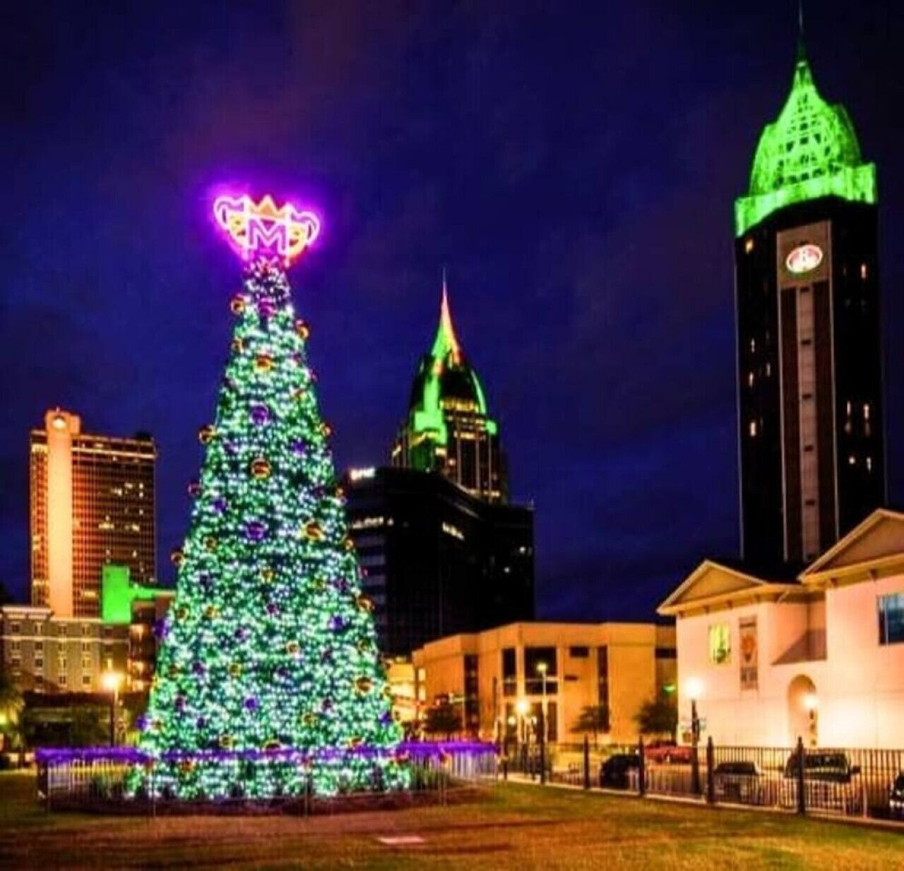 A large Christmas tree with lights and a neon sign at the top, surrounded by buildings and a clock tower.