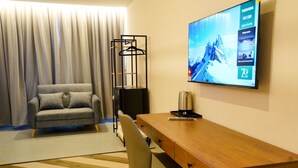 Une chambre d’hôtel moderne avec un canapé gris, un bureau en bois et un téléviseur fixé au mur qui diffuse une image de paysage montagneux.
